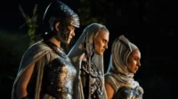 Galadriel em armadura élfica e duas figuras encapuzadas de Os Anéis de Poder olham à frente em cenário noturno, com ar sério.