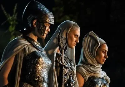 Galadriel em armadura élfica e duas figuras encapuzadas de Os Anéis de Poder olham à frente em cenário noturno, com ar sério.