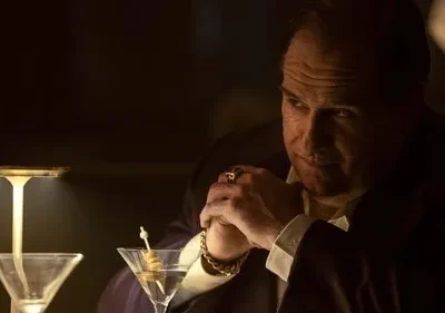 Homem de terno, com anel e pulseira, mãos postas sobre a mesa, olha pensativo em um bar escuro. Dois martínis estão na frente.