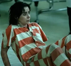 Jovem de uniforme prisional laranja e branco, sentada no chão de um ambiente austero. Ela olha apreensiva para o lado.