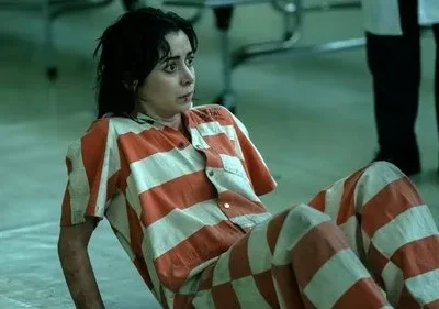 Jovem de uniforme prisional laranja e branco, sentada no chão de um ambiente austero. Ela olha apreensiva para o lado.