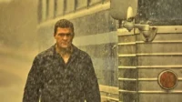 Pôster de Reacher. Alan Ritchson, encharcado pela chuva, olha intensamente para frente, com um ônibus azul e prateado ao fundo. Clima tenso.