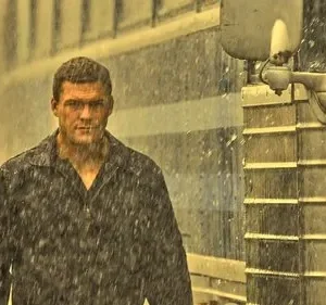 Pôster de Reacher. Alan Ritchson, encharcado pela chuva, olha intensamente para frente, com um ônibus azul e prateado ao fundo. Clima tenso.