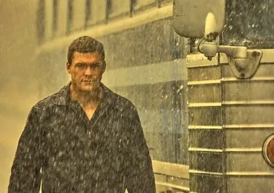 Pôster de Reacher. Alan Ritchson, encharcado pela chuva, olha intensamente para frente, com um ônibus azul e prateado ao fundo. Clima tenso.