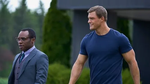 Da série Reacher:Alan Ritchson (musculoso,camiseta azul) e Malcolm Goodwin (terno,óculos) caminham ao ar livre. Clima de ação e investigação.