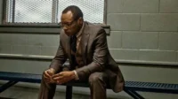 Homem de terno e óculos, de When They See Us, sentado pensativo num banco de metal em ambiente com parede cinza e grade.