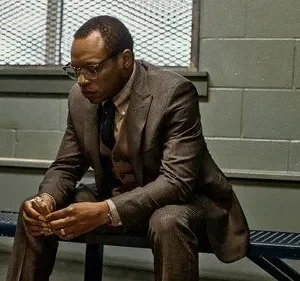 Homem de terno e óculos, de When They See Us, sentado pensativo num banco de metal em ambiente com parede cinza e grade.