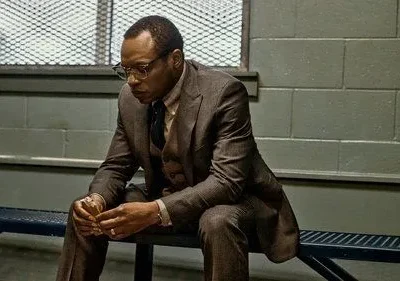 Homem de terno e óculos, de When They See Us, sentado pensativo num banco de metal em ambiente com parede cinza e grade.
