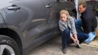 Em Reacher, uma mulher tensa de jaqueta cinza com arma e um homem agacham-se atrás de um SUV cinza em cena de perigo.