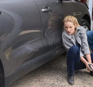 Em Reacher, uma mulher tensa de jaqueta cinza com arma e um homem agacham-se atrás de um SUV cinza em cena de perigo.