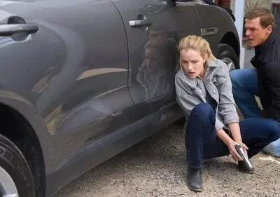 Em Reacher, uma mulher tensa de jaqueta cinza com arma e um homem agacham-se atrás de um SUV cinza em cena de perigo.