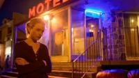 Julia Garner (Ruth) em Ozark, pensativa, de braços cruzados, à noite. Um motel com letreiro neon vermelho e luz azul cria um clima dramático.