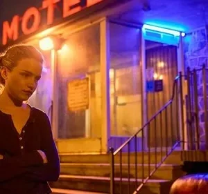 Julia Garner (Ruth) em Ozark, pensativa, de braços cruzados, à noite. Um motel com letreiro neon vermelho e luz azul cria um clima dramático.