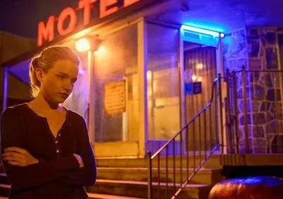 Julia Garner (Ruth) em Ozark, pensativa, de braços cruzados, à noite. Um motel com letreiro neon vermelho e luz azul cria um clima dramático.