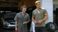 Reacher, musculoso, paga mecânico sorridente Dale em oficina de carros com capô aberto. Cena da série Reacher.
