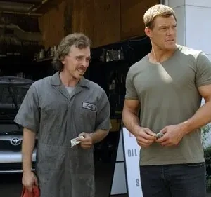 Reacher, musculoso, paga mecânico sorridente Dale em oficina de carros com capô aberto. Cena da série Reacher.