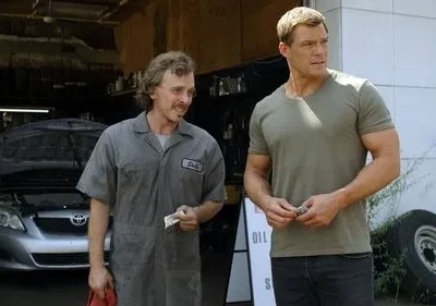 Reacher, musculoso, paga mecânico sorridente Dale em oficina de carros com capô aberto. Cena da série Reacher.