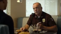 Em Fargo, um policial de uniforme marrom com distintivo senta em um diner comendo um lanche, conversando seriamente com alguém.