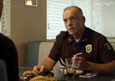 Em Fargo, um policial de uniforme marrom com distintivo senta em um diner comendo um lanche, conversando seriamente com alguém.
