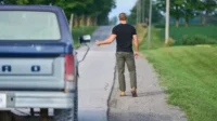 Em O Fugitivo, um homem musculoso de camiseta preta e calça verde caminha por estrada rural, pedindo carona. Caminhão Ford azul parado à esquerda.
