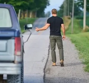 Em O Fugitivo, um homem musculoso de camiseta preta e calça verde caminha por estrada rural, pedindo carona. Caminhão Ford azul parado à esquerda.