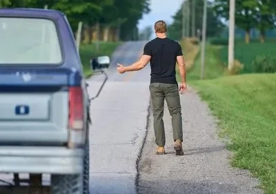 Em O Fugitivo, um homem musculoso de camiseta preta e calça verde caminha por estrada rural, pedindo carona. Caminhão Ford azul parado à esquerda.