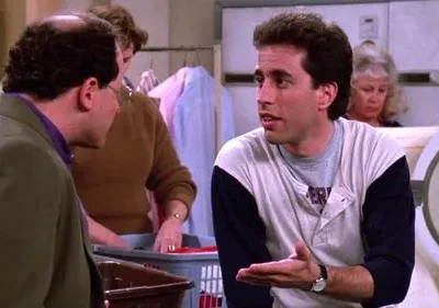 Jerry Seinfeld gesticula enquanto conversa com George Costanza em uma lavanderia. Típica cena da série Seinfeld.