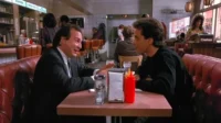 Em um diner clássico de Seinfeld, Jerry Seinfeld ouve George Costanza, que gesticula animadamente em uma cabine vermelha, em um clima cômico.