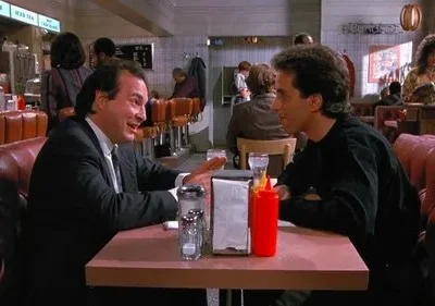 Em um diner clássico de Seinfeld, Jerry Seinfeld ouve George Costanza, que gesticula animadamente em uma cabine vermelha, em um clima cômico.