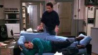 Jerry Seinfeld gesticula, exasperado, enquanto George Costanza cochila no sofá com sapatos, em uma cena clássica de Seinfeld.