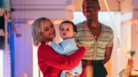 Doctor Who – T01E01:Bebês do Espaço 10 Cena de série/filme:Mulher loira sorridente segura bebê no colo,enquanto um homem de camisa listrada sorri ao fundo. Ambiente com luzes coloridas.