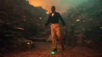 Doctor Who – T01E03:Boom 8 Homem elegante flutua em plataforma verde num cenário rochoso com fogo,exalando um clima de aventura sci-fi. Pôster de série/filme.