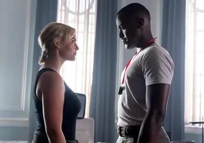 Cena de [Nome da Série/Filme]: mulher loira e homem negro trocam olhares tensos em um elegante cômodo com janelas grandes.