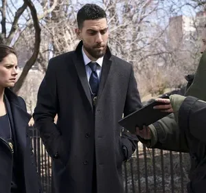 Os agentes do FBI Maggie e OA, apreensivos, observam um técnico em traje antibomba com um tablet em cena urbana.