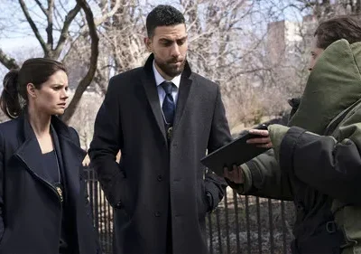 Os agentes do FBI Maggie e OA, apreensivos, observam um técnico em traje antibomba com um tablet em cena urbana.