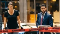 Pôster de FBI:Agentes sérios Maggie e Omar,atrás de fita HAZARDOUS MATERIAL em cena urbana,com clima tenso.