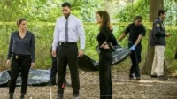 Personagens de Criminal Minds:Prentiss,Simmons e Lewis em cena de crime na floresta,com um corpo sendo removido. (115 caracteres)