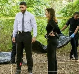 Personagens de Criminal Minds: Prentiss, Simmons e Lewis em cena de crime na floresta, com um corpo sendo removido. (115 caracteres)