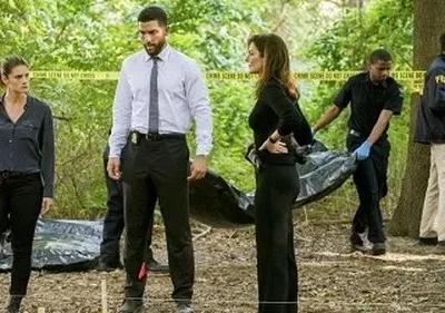 Personagens de Criminal Minds: Prentiss, Simmons e Lewis em cena de crime na floresta, com um corpo sendo removido. (115 caracteres)