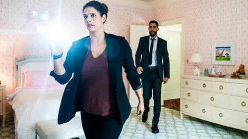 Em FBI,uma detetive com lanterna explora um quarto infantil rosa. Um agente de terno entra. Ambiente tenso de investigação.