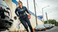 Agente do FBI correndo com colete tático em rua urbana. Carros e edifícios ao fundo em cena de ação da série FBI.