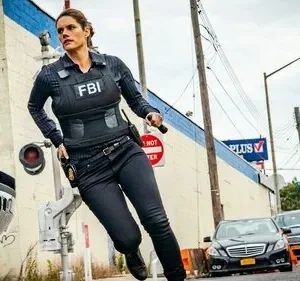 Agente do FBI correndo com colete tático em rua urbana. Carros e edifícios ao fundo em cena de ação da série FBI.