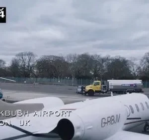 Cena de série: Um jato particular branco em Blackbush Airport, Londres, UK, com um funcionário e veículos de apoio sob céu nublado.