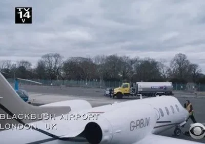 Cena de série: Um jato particular branco em Blackbush Airport, Londres, UK, com um funcionário e veículos de apoio sob céu nublado.