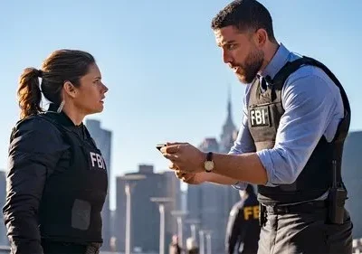 Agentes do FBI, uma mulher e um homem em coletes táticos, conversam seriamente em cenário urbano ensolarado, da série FBI.