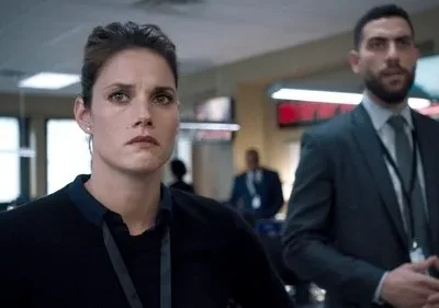 Mulher séria em primeiro plano e homem ao fundo em escritório tenso, com monitores. Imagem capta a urgência de uma série/filme de suspense.