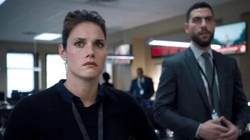 FBI – T01E14:Denúncia 1 Mulher séria em primeiro plano e homem ao fundo em escritório tenso,com monitores. Imagem capta a urgência de uma série/filme de suspense.