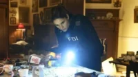A agente de FBI está concentrada,iluminando uma mesa com objetos diversos. O clima é de investigação tensa na série FBI.