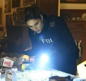 A agente de FBI está concentrada, iluminando uma mesa com objetos diversos. O clima é de investigação tensa na série FBI.
