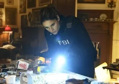 A agente de FBI está concentrada, iluminando uma mesa com objetos diversos. O clima é de investigação tensa na série FBI.